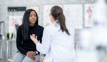 A woman of African descent and her doctor are indoors in a medical clinic. They are sitting and talking about the woman's health problems.